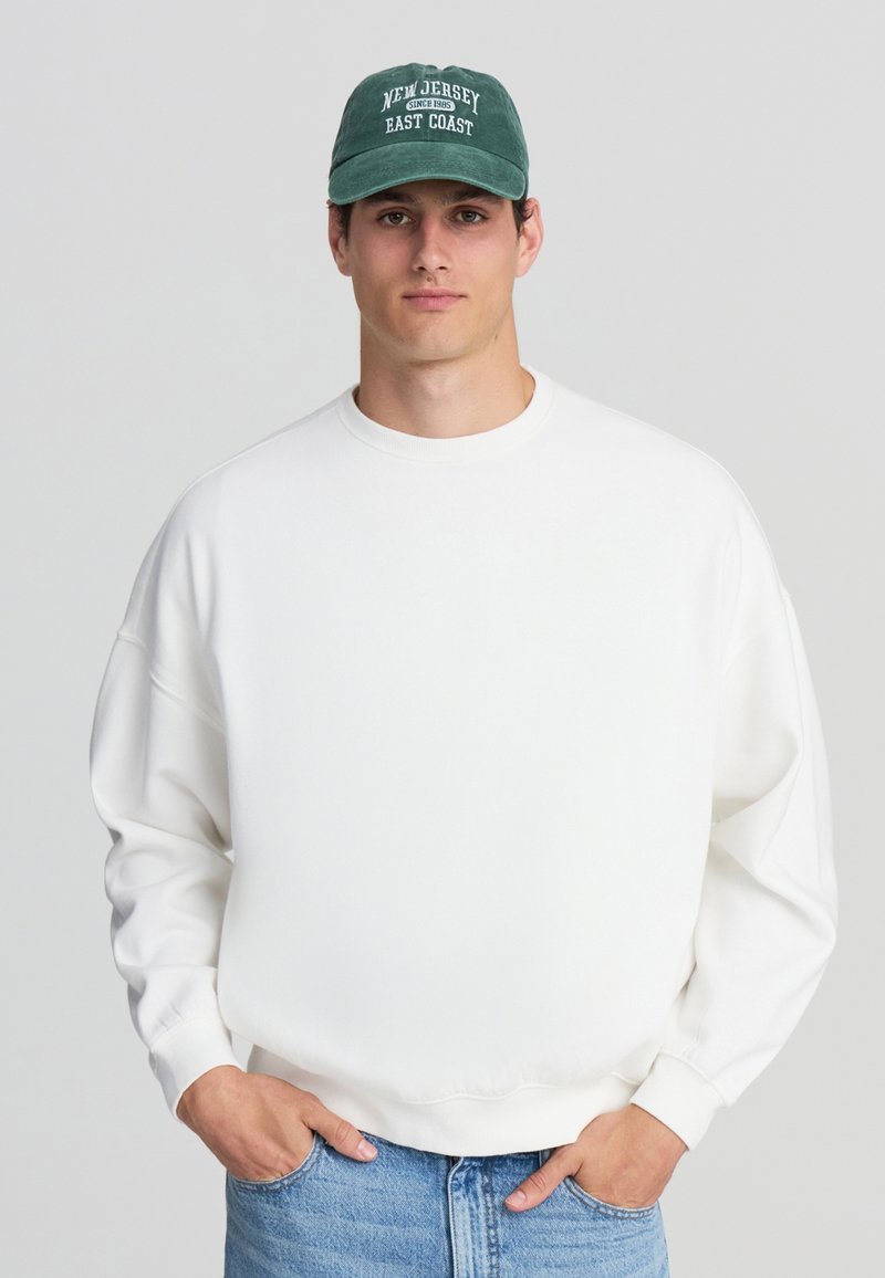 Felpa bianca oversize con scollo tondo e maniche a sbuffo, abbinata a un berretto verde scuro con testo bianco. Jeans in denim.