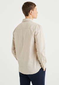 Light beige long-sleeve shirt made of textured linen with a collar and buttoned cuffs, featuring a rounded hem and back yoke detail.