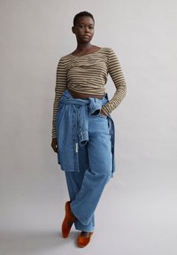 Gestreiftes langärmliges Oberteil in Braun und Beige, kombiniert mit lockeren blauen Jeans. Das Model trägt orangefarbene High Heels und bindet ein hellblaues Jeanshemd um die Taille.