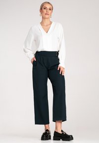 Biała bluzka z dekoltem w serek i długimi rękawami, w połączeniu z ciemnozielonymi spodniami szerokimi w nogawkach z detalem guzika oraz czarnymi masywnymi loafersami.