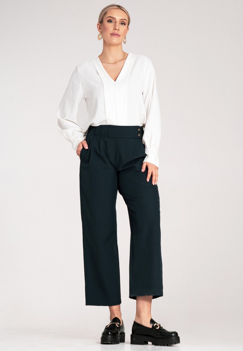 Biała bluzka z dekoltem w serek i długimi rękawami, w połączeniu z ciemnozielonymi spodniami szerokimi w nogawkach z detalem guzika oraz czarnymi masywnymi loafersami.