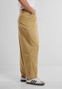 Beige, straight-leg trousers made of cotton with a casual design. Features side pockets and a back pocket, paired with white sneakers.