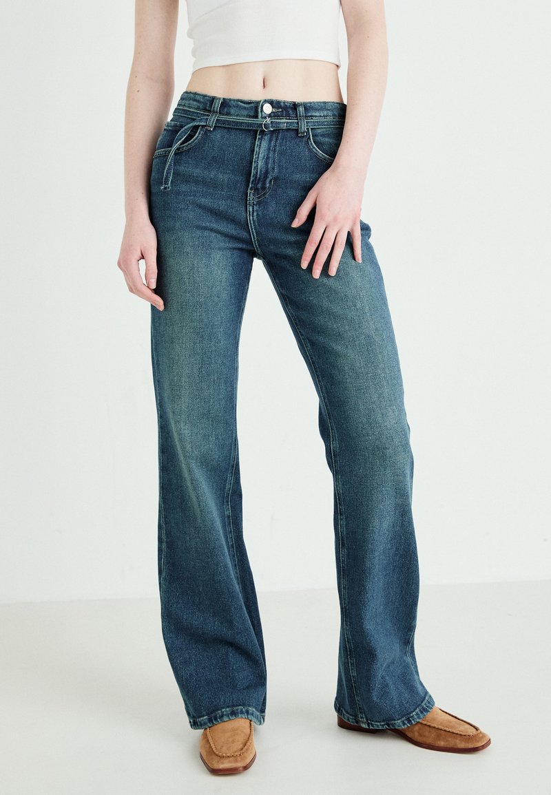Jeans a zampa d'elefante in denim blu con vita alta, caratterizzati da tasche frontali, chiusura bottone e un aspetto sbiadito. Indossati con mocassini marroni.