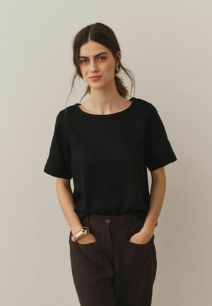 Schwarzes T-Shirt mit kurzen Ärmeln und rundem Halsausschnitt, aus glattem Stoff, kombiniert mit braunen Hosen mit sichtbaren Taschen. Einfaches Design ohne Muster.