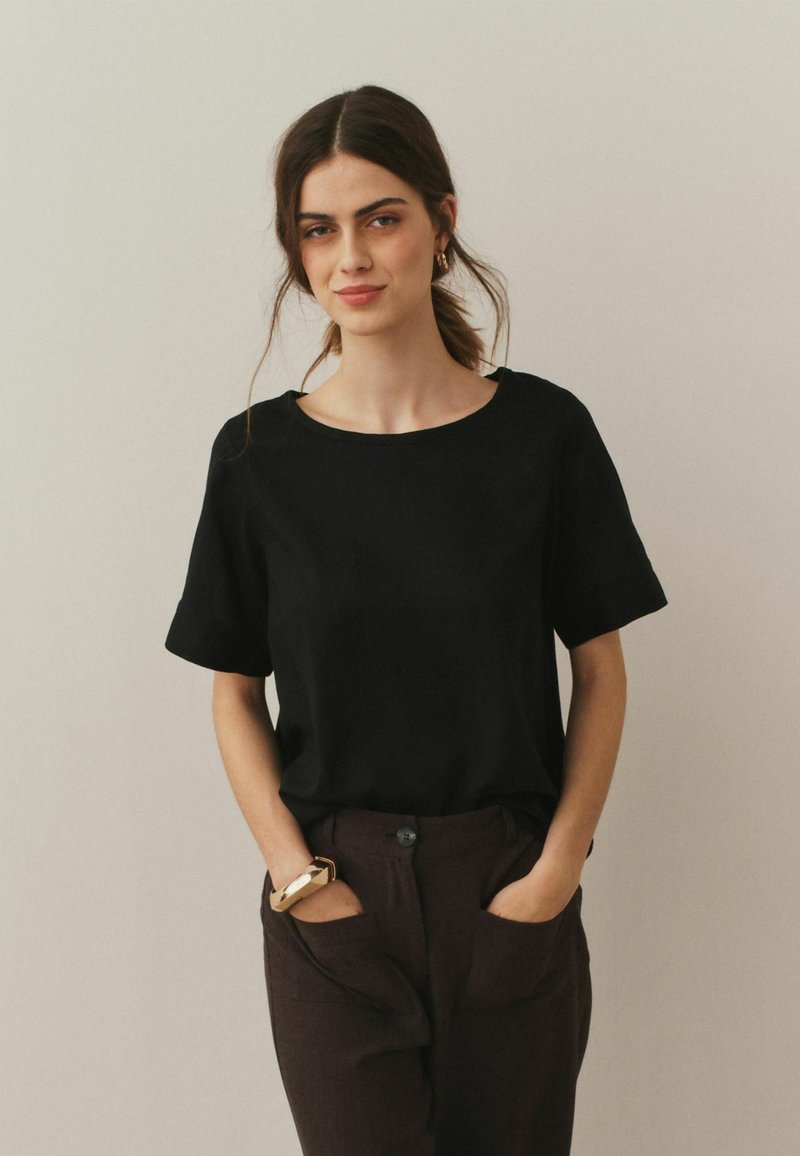 T-shirt noir à manches courtes avec un col rond, fabriqué en tissu lisse, associé à un pantalon marron avec poches apparentes. Design simple sans motifs.