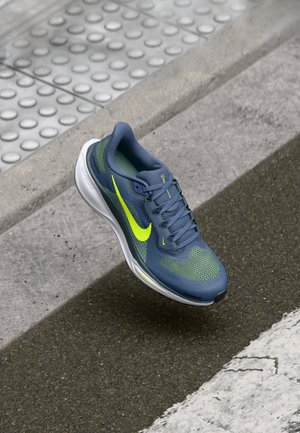 Niebieski i neonowo zielony but do biegania Nike unoszący się nad asfaltową drogą w pobliżu powierzchni dotykowej i białego oznakowania drogowego.