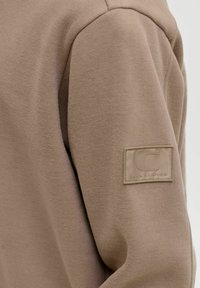 Beige fliisist valmistatud jope varrukas on õmmeldud ristkülikukujuline ilmuv patch, millel on tõstetud "C" ja "JACK & JONES" tekst.
