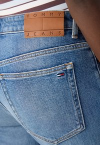 Lys blå denimjeans med en brun lærpatch merket "TOMMY JEANS" over høyre baklomme og en liten rød, hvit og blå logo.