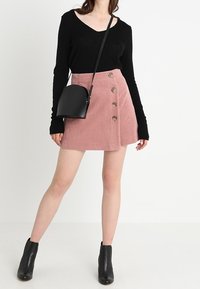 Maglione nero a maniche lunghe abbinato a una gonna di velluto a coste rosa con bottoni davanti. Una borsa a tracolla nera e stivaletti alla caviglia completano l'outfit.
