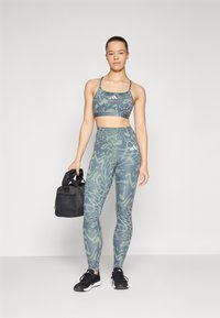 Sutiã desportivo e leggings em tecido verde com padrão. As leggings têm uma cintura alta e detalhes brancos subtis. O modelo segura uma mala de ginásio preta.