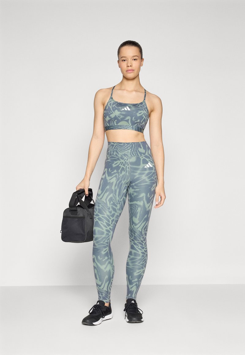 Sujetador deportivo y leggings de tela verde con estampado. Los leggings tienen una cintura alta y sutiles acentos blancos. La modelo sostiene un bolso de gimnasio negro.