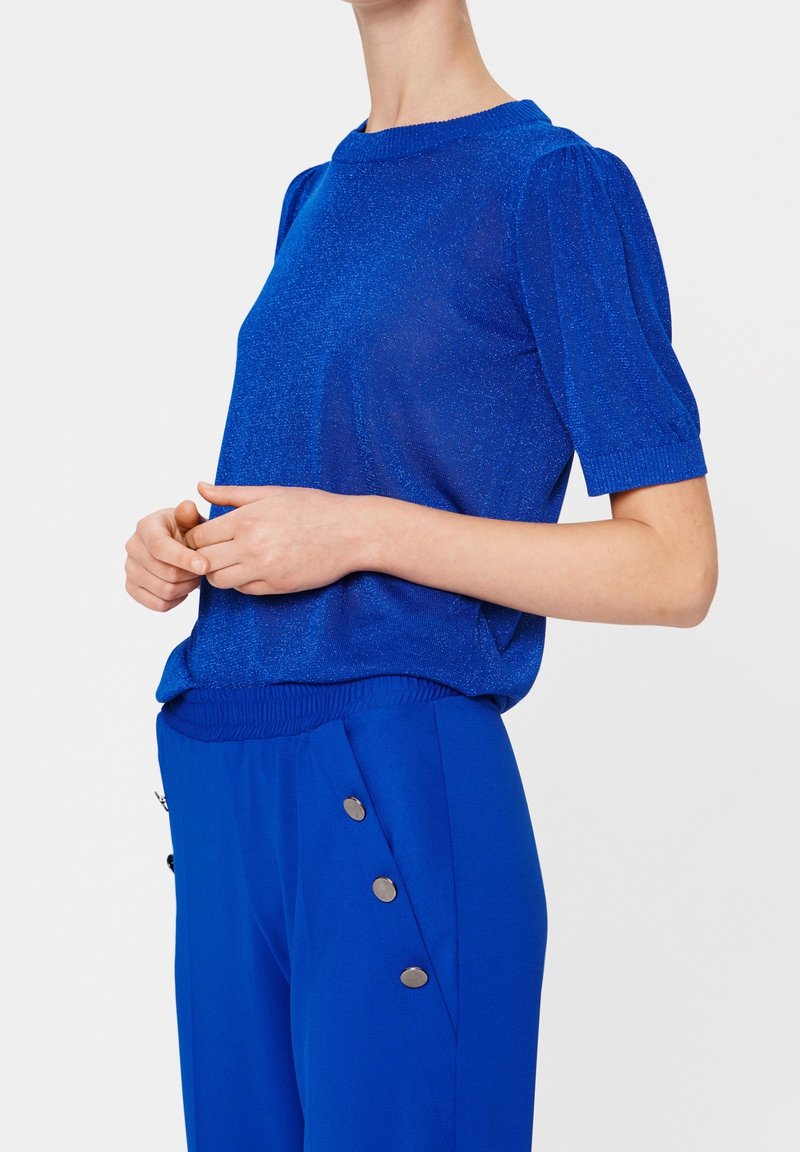 Femme portant un haut bleu vif à manches courtes bouffantes et un pantalon taille haute assorti avec trois boutons argentés sur le côté.