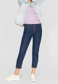 Jeans dżinsowe w ciemnoniebieskim kolorze o kroju zwężanym, w połączeniu z jasnofioletową bluzką i jasnoniebieską kurtką dżinsową. Białe sneakersy sportowe.