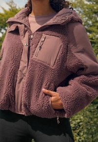 Osoba ubrana w fioletową polarową kurtkę z teksturowanej tkaniny i kieszeniami na zamek, stojąca na zewnątrz z drzewami w tle.