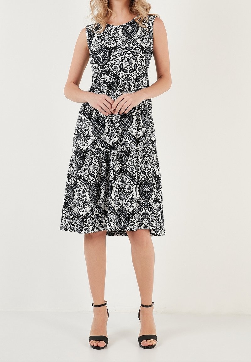 Femme portant une robe sans manches noire et blanche à motif, longueur genoux, avec des talons hauts noirs à bride cheville, debout devant un fond uni.