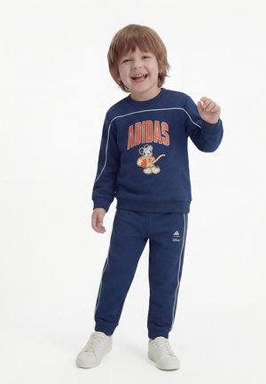 Naeratav noor poiss, kes kannab tumesinist Adidas Disney dressipluusi, millel on Mickey Mouse'i kujutis, seisab ja tõstab ühe käe valgel taustal.