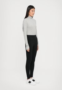 Grijze langwerpige fitted top met hoge kraag, gecombineerd met zwarte leggings. Het model draagt witte sneakers met zwarte accenten. Glad textiel met een soepele textuur.