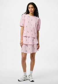 Completo due pezzi rosa con top a maniche corte e gonna stratificata, con dettagli di ricamo floreale. Indossato con scarpe sportive bianche.