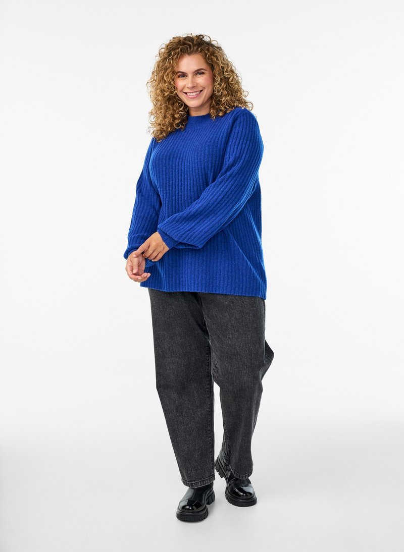 Smilende kvinde med krøllet hår iført en blå sweater, sorte jeans og sorte sko, stående mod en hvid baggrund.