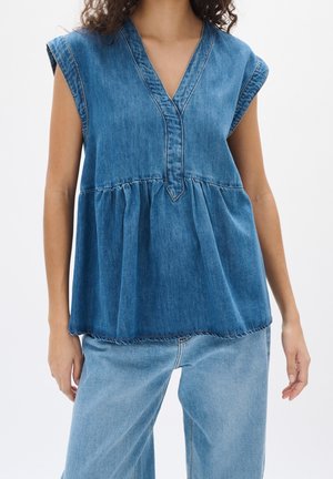 Kvinde iført ærmeløs blå denimtop med V-hals og rynket talje, kombineret med lyseblå denimjeans.