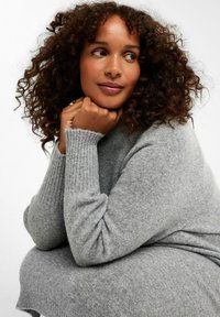 Hellgrauer Strickpullover mit gerippten Bündchen und glatter Textur. Das Model sitzt und stützt das Kinn auf die Hand, hat lockige Haare und einen neutralen Hintergrund.