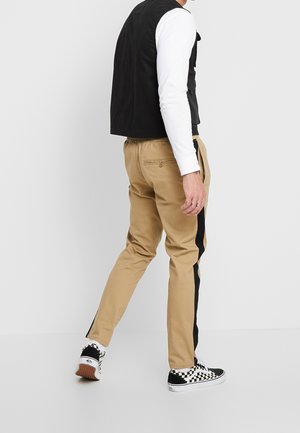 Osoba w beżowych spodniach z czarnymi bocznymi paskami, czarno-białych tenisówkach w kratę, białej koszuli z długim rękawem i czarnej kamizelce, stojąca bokiem.