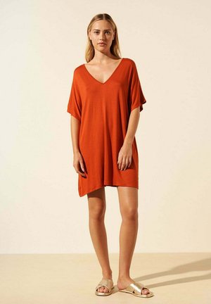 Robe orange en tissu avec un décolleté en V et des manches courtes, tombant juste au-dessus des genoux. Le mannequin porte des sandales à glisser dorées. Fond uni.