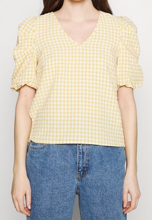 Mujer con blusa de cuadros amarillos y blancos con escote en V y mangas cortas abullonadas, combinada con vaqueros azules.
