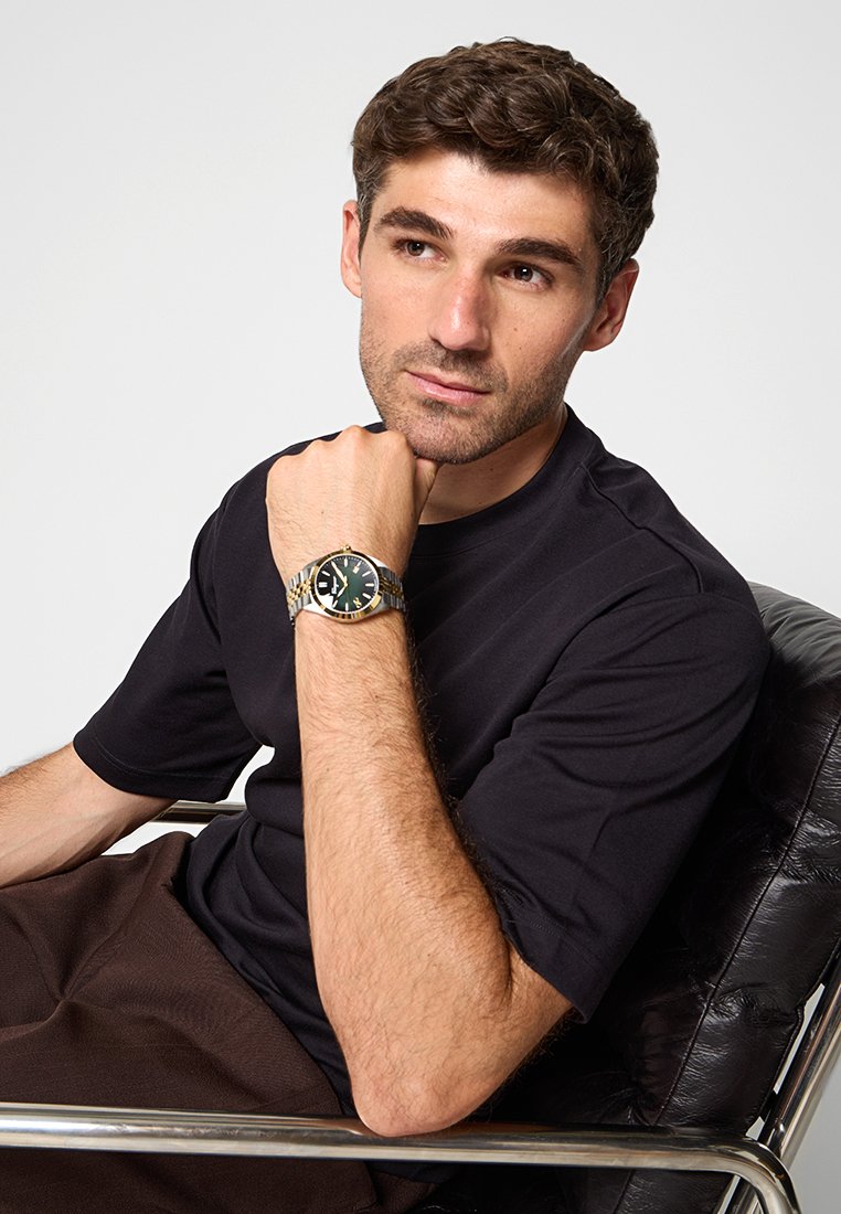 Montre-bracelet argentée et dorée avec un cadran vert, portant des index horaires numériques. Le modèle porte un t-shirt noir à col rond et est assis sur une chaise noire.