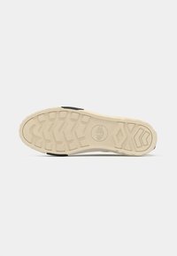 Λάστιχο πάτου αθλητικού παπουτσιού με μπεζ εξωτερικό στρώμα, υφή πέλματος με σχέδιο και κυκλικό λογότυπο στο κέντρο, με μαύρη λεπτομέρεια.