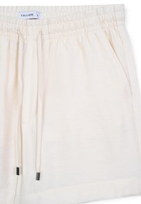 Shorts beige clair en tissu doux, dotés d'une taille élastique, d'un cordon de serrage avec des embouts en métal et de poches latérales. Texture lisse.