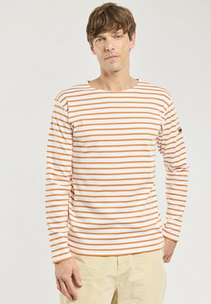 Homme portant une chemise blanche à manches longues rayée horizontalement orange et un pantalon beige, debout devant un fond clair uni.