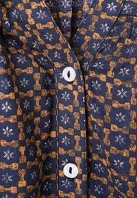 Mønstret skjorte i marineblå og sennep, prydet med et gitterdesign og blomster- samt vægte-motiver. Den har hvide knapper og en tekstureret overflade.