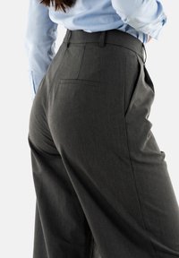 Pantaloni tailored grigio scuro con vita alta, pieghe e tasca posteriore. Il tessuto ha una texture liscia, abbinati a una camicia azzurro chiaro.