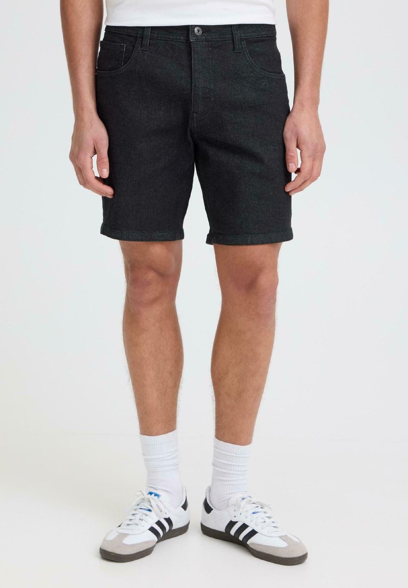 Homme portant un short en jean noir, des chaussettes blanches montantes et des baskets blanches avec des rayures noires, debout devant un fond clair uni.