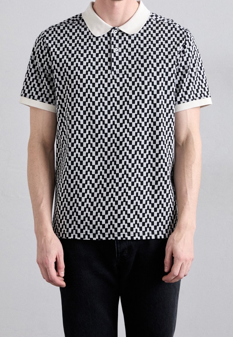 Homme portant un polo à manches courtes avec motif géométrique noir et blanc, col blanc et poignets blancs, associé à un pantalon noir.