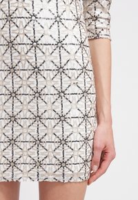 Robe blanche avec motifs géométriques en sequins noirs et beiges, manches trois-quarts et silhouette ajustée, montrée de la mi-cuisse au poignet.
