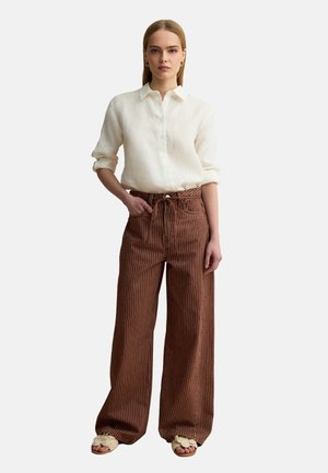 Woman wearing a white button-up shirt, brown wide-leg pinstriped pants with a tie waist, and beige braided open-toe sandals, standing with one hand in pocket.