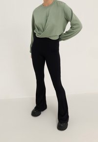 Persona vistiendo una camiseta de manga larga verde claro y pantalones acampanados negros de cintura alta con zapatos gruesos negros, de pie contra un fondo claro y liso.