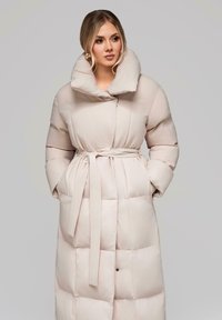 Žena nosící dlouhý, světlobéžový prošívaný kabát s širokým límcem a uvázaným páskem, s rukama v kapsách, dívající se stranou.