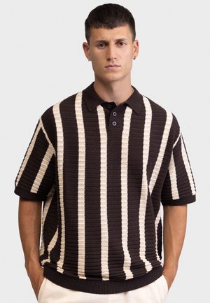 Νέος άνδρας με κοντά σκούρα μαλλιά, φοράει κοντομάνικο πλεκτό πόλο μπλουζάκι με κάθετες μαύρες και άσπρες ρίγες, στέκεται με τα χέρια στις τσέπες ενός ανοιχτόχρωμου παντελονιού.