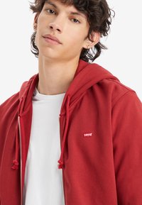 Giovane con capelli corti e ricci che indossa una felpa rossa zip-up di Levi's sopra una maglietta bianca semplice su uno sfondo bianco.