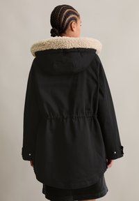 Zwarte parka jas met een zachte roomkleurige kraag, elastische tailleband en knoopmanchetten, gemaakt van een gladde buitenstof.