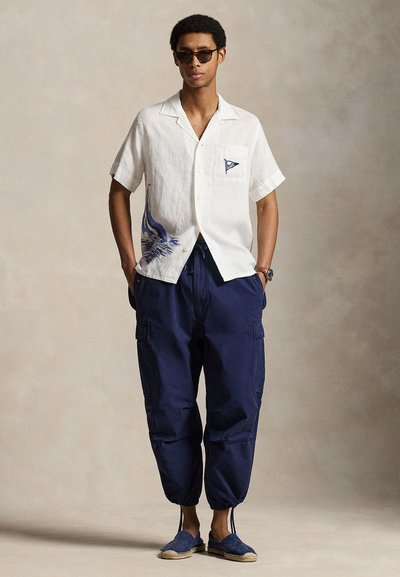 Homme portant une chemise blanche à manches courtes avec un motif bleu, un pantalon cargo bleu marine avec des ourlets à cordon, des lunettes de soleil et des espadrilles bleues, debout.