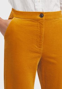 Rippstoffhosen in einer satten Senffarbe mit strukturiertem Oberflächenmaterial, ausgestattet mit einem Frontknopfverschluss und seitlichen Taschen.