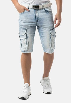 Person wearing light blue denim cargo shorts, white belt, white sneakers, standing with one hand in pocket against plain background.