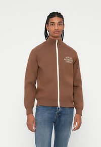 Brun genser med glidelås og høy krage, hvit glidelås, og logo-tekst på venstre bryst. Fitted fasong med ribbede mansjetter og nederkant. Brukt sammen med blå jeans.