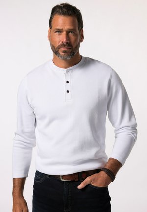 Camisa blanca de manga larga estilo henley con tres botones oscuros en la tapeta, textura acanalada, combinada con jeans oscuros y un cinturón marrón.