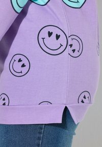 Paarse katoenen sweatshirt met zwarte smiley-face patronen en een zijsplitontwerp, gestopt in een blauwe spijkerbroek.