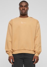 Beige sweatshirt med rund halsudskæring, rib strik ved håndleddene og frynset kant. Har et hvidt broderet logo på tværs af brystet. Normal pasform.